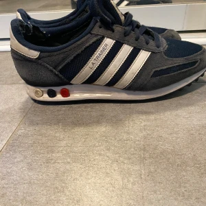Adidas LA Trainer sneakers blå/vit - Snygga Adidas LA Trainer sneakers i marinblått och vitt med klassiska tre ränder på sidan. Ovandel i mesh och mocka, snörning och cool detalj med färgade pluggar i sulan. Perfekta skor inför vintern!