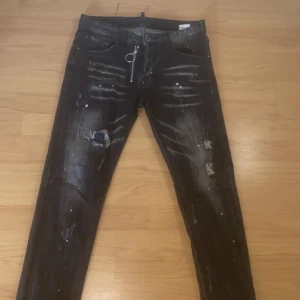 Svarta dsquared2 jeans  - Säljer ett par svarta jeans från Dsquared2 med slitna detaljer, vit färgskvätt och cool dragkedja med ring. Jeansen har en tvättad look och är tillverkade i stretchig bomull för en tajt passform. Perfekta för dig som gillar streetstyle.