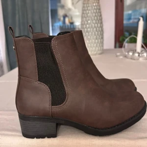 Bruna chelsea boots från Clou - Snygga bruna chelsea boots från Clou i storlek 38. Skorna har elastiska svarta paneler på sidorna och en låg klack. Tillverkade i ett material som liknar skinn och har rund tå. Perfekta för höst och vinter med sin klassiska look.