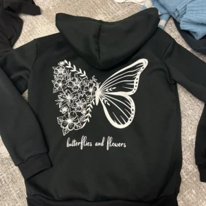 Svart hoodie med fjäril och blommor - Svart hoodie med stor vit print på ryggen som föreställer en fjäril där ena vingen är fylld med blommor och blad. Texten 'butterflies and flowers' under motivet. Klassisk huva och magficka framtill. Perfekt för dig som gillar unika tryck och naturinspirerade detaljer.