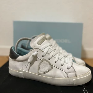 Vita sneakers från Philippe Model - Snygga vita sneakers från Philippe Model med grå detalj på sidan och svart häl. Skorna har rund tå, platt sula och klassisk snörning. Tillverkade i skinn för en lyxig känsla och stilren look. Perfekta för dig som gillar minimalistisk design. Mycket bra skick! Storlek 40 passar också 41. Hör av er ifall ni har några frågor!