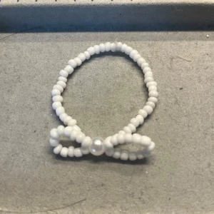 Pärlarmband med rosett i vitt - Söt armband gjord av små vita pärlor, formad som en rosett framtill med en lite större pärla i mitten. Perfekt accessoar för att ge en gullig touch till din stil. Handgjort!