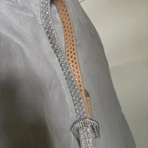 Glittrigt silverbälte med strass - Säljer ett snyggt bälte i silver med massor av glittrande strass och nitar. Bältet har en klassisk form och spänne som också är täckt av stenar. Insidan är beige och bältet är gjort i ett material som liknar skinn. Perfekt för att lyfta vilken outfit som helst!