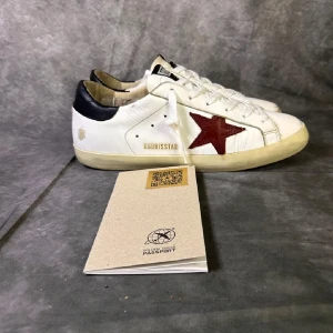 Golden Goose vita sneakers med röd stjärna - Golden Goose sneakers i vitt skinn med ikonisk röd stjärna på sidan och svarta detaljer vid hälen. Klassisk låg modell med snörning och platt sula. Snygg vintage-look och coola kontraster som sticker ut.