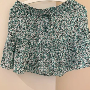 Blommig kortkjol med volang och knyt - Supersöt kortkjol med blommigt mönster i turkos, svart och vitt. Kjolen har volangkant och en liten knytning i midjan. Perfekt för dig som gillar en lätt och flowy stil till sommaren.