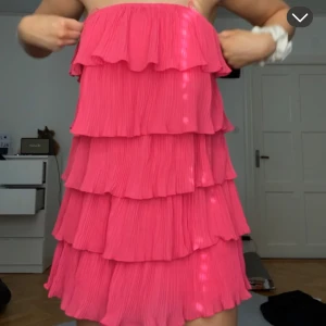 Axelbandslös rosa volangklänning - Superfin axelbandslös kortklänning i starkt rosa med flera lager volanger. Klänningen har band dock som man kan ha över axlarna men jag har använt den som bandau. Går att ha båda. Använt några få tal gånger. Jag är 174