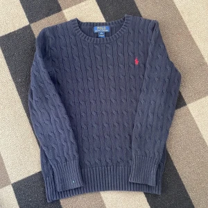 Mörkblå stickad tröja Polo Ralph Lauren - Kabelstickad mörkblå tröja från Polo Ralph Lauren med klassisk rund hals och röd broderad logga på bröstet. Tröjan är långärmad och har ribbade muddar och nederkant. Perfekt för dig som gillar preppy och stilren look. Priset ej hugget i sten