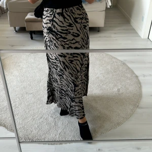 Beige långkjol med zebramönster - Säljer en beige långkjol med svart zebramönster. Kjolen har en flowy passform och är tillverkad i ett mjukt, lätt material som ger snyggt fall. Perfekt för dig som vill sticka ut med ett djurmönster och ändå hålla det stilrent.