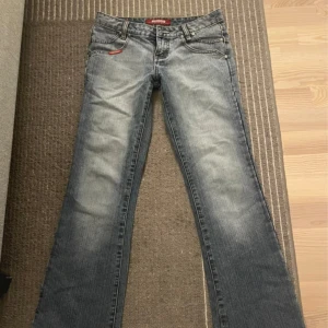 Grå lågmidjade bootcut jeans Venus Ross - Snygga grå bootcut jeans från Venus Ross med slitningar och coola broderade detaljer på bakfickorna. Klassisk femficksmodell med orange logotyp på linningen. Perfekta för dig som gillar en avslappnad men trendig stil.