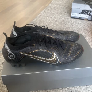 Nike Vapor 14 Elite AG fotbollsskor - Nike Vapor 14 Elite AG fotbollsskor i svart med metallicguld-detaljer. Skorna har snörning, låg profil och en ovandel i syntetmaterial med textur för extra grepp. Snygg design med guldiga linjer och silverdetaljer på hälen. Perfekta för spel på konstgräs.