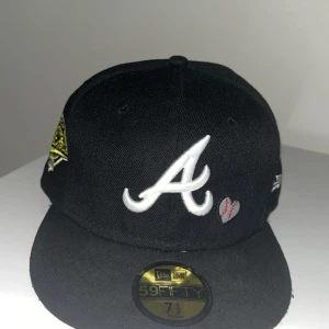 Svart Atlanta Braves keps New Era - Svart 59FIFTY keps från New Era med Atlanta Braves A-logga framtill, broderat hjärta med baseboll-sömmar och texten 'Love & Hustle = Game' bak. MLB-logga bak och gul detalj på sidan. Klassisk flat peak och broderade detaljer.storlek 59,6cm