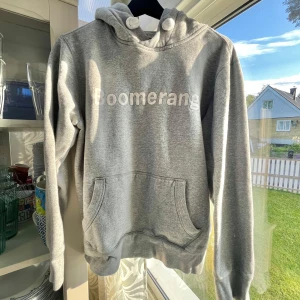 Grå hoodie från Boomerang med huva - Snygg grå hoodie från Boomerang med vit logga framtill. Tröjan har stor huva med grova snören och en klassisk känguruficka. Mjuk insida och ribbade muddar vid ärmslut och nederkant. Perfekt för chill dagar eller när du vill ha en enkel och stilren look.