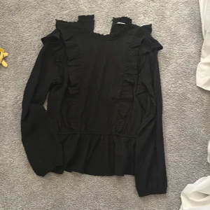 Svart blus med volangdetaljer - Säljer en svart blus med långa ärmar och snygga volangdetaljer längs axlar och framsida. Blusen har en feminin och trendig look, perfekt att styla med jeans eller kjol. Materialet känns lätt och luftigt, vilket gör den skön att bära.
