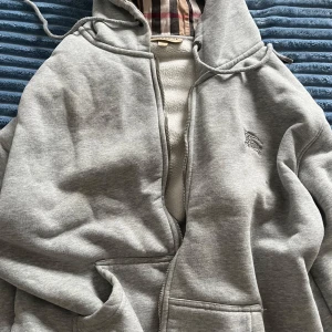 Grå hoodie från Burberry med fleece - Säljer en grå hoodie från Burberry i storlek XL men passar L och M. Utsidan är i mjuk  och insidan har ett varmt fleecefoder. Klassisk Burberry-rutig huva, dragkedja framtill och broderad logga på bröstet. Perfekt för dig som gillar stilren och lyxig streetwear.