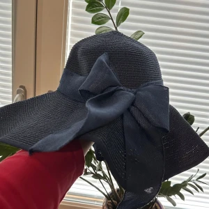 Svart solhatt med stor rosett - Snygg svart solhatt med bred brätte och en stor dekorativ rosett på sidan. Hatten är flätad och har en klassisk form som ger skön skugga. Perfekt för soliga dagar och ger en chic vibe till din outfit.