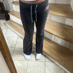 Juicy Couture mörk grå mjukisbyxor - Mörkgråa mjukisbyxor från Juicy Couture med  logga och strassdetaljer på ena benet. Byxorna har snörning i midjan och är tillverkade i mjuk velour. Klassisk bootcut-modell med låg midja som ger en avslappnad men ändå snygg look. Finns några diskreta klorin fläckar.