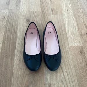 Svarta ballerinaskor från H&M - Klassiska svarta ballerinaskor från H&M med rund tå och söt rosettdetalj framtill. Skorna har en platt sula. Perfekta för en stilren och enkel look. Använda en gång men säljer pågrund av att dem var för stora. Inga speciella defekter, fått lite på ena framsida tån.