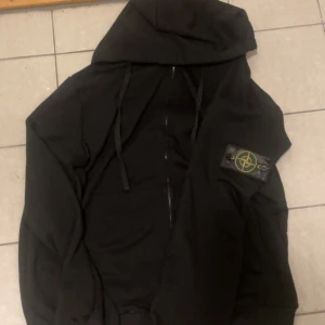 Svart hoodie från Stone Island - Svart hoodie från Stone Island med dragkedja och klassisk patch på ärmen. Hoddien har huva med snörning och är tillverkad i mjukt bomullsmaterial. Perfekt för en chill och stilren look.