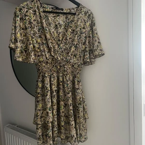 Mönstrad kortklänning från Zara - Supersöt kortklänning från Zara med v-ringning, smockad midja och volangig kjol. Klänningen har ett färgstarkt abstrakt mönster i beige, svart, grön och gul. Lätt och luftig med korta ärmar, perfekt för varma dagar. 