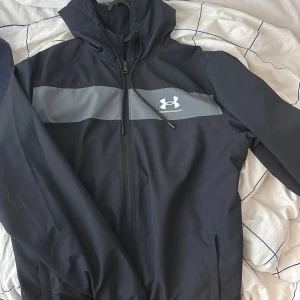 Svart vindjacka från Under Armour - Säljer en svart vindjacka från Under Armour med grått band över bröstet och dragkedja framtill. Jackan har en hög krage och långa ärmar, perfekt för blåsiga dagar. Den är designad för en loose passform och har det ikoniska Under Armour-logotypen på bröstet. M i storlek men passar även M