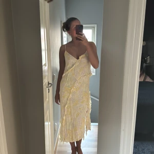 Blommig sommarklänning med volang - Superfin ljus klänning med blommigt mönster i gult, grönt och lila. Den har smala axelband, v-ringning och volangdetaljer som går snett över klänningen. Lätt och flowy känsla, perfekt för varma dagar.