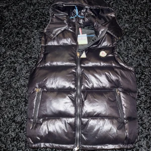 Svart dunväst från Moncler med huva - Säljer en svart, glansig dunväst från Moncler med avtagbar huva och dragkedja framtill. Västen har två fickor med dragkedja och klassisk Moncler-logga på bröstet. Perfekt för lager-på-lager och riktigt snygg till streetwear-stilen.
