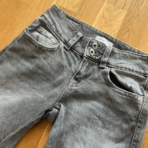 Grå bootcut jeans med dubbla knappar - Snygga grå jeans från med bootcut-modell och låg midja. Jeansen har dubbla knappar i midjan, klassiska fem fickor och bakfickor med lock och knapp. Tillverkade i mjukt denimtyg som ger en skön känsla och cool look.