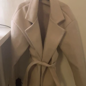 Beige kappa med bälte och bred krage - Snygg beige kappa med breda slag och knytskärp i midjan. Jackan har lång ärm och en klassisk, stilren look som passar perfekt till höst och vinter. Materialet ser ut att vara ullblandning för en mjuk och varm känsla.