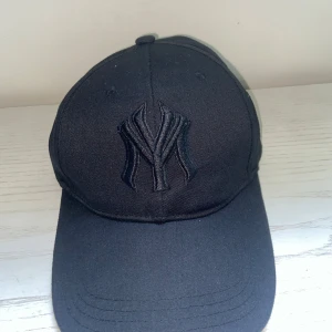 Svart New York Yankees keps - Snygg svart keps med böjd skärm och broderad NY-logga framtill. Justerbar rem baktill med metallspänne. Klassisk basebollstil, perfekt för en sportig look. Tillverkad i bomullstyg för skön passform.