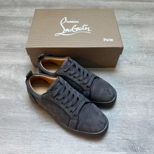 Mörkgrå sneakers från Christian Louboutin - Snygga mörkgrå sneakers från Christian Louboutin i mocka med klassisk rund tå och platt sula. Skorna har breda snören och den ikoniska röda sulan med Louboutin-logga. Perfekta för dig som vill ha en stilren och lyxig look.