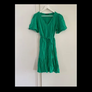 Grön kortklänning - Superfin grön kortklänning från Papaya med v-ringning, korta puffärmar och knytband i midjan. Klänningen har en luftig känsla och är gjord i 100% muslin bomull, perfekt för varma dagar. Den har en lätt volang nertill för extra flow. Längd 106cm. Storleken passar från 42 till 46.