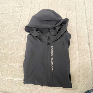 Svart vindjacka från Under Armour - Svart vindjacka från Under Armour med huva och hel dragkedja framtill. Jackan har diskret logga på bröstet och är tillverkad i lätt polyester med meshfoder. Perfekt för träning utomhus och har två fickor med dragkedja.