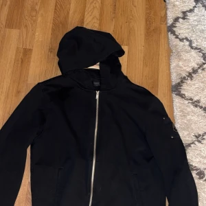 Svart vindjacka från Moose Knuckles - Svart vindjacka från Moose Knuckles med huva och hel dragkedja framtill. Jackan har en snygg metallogo på ärmen och praktiska fickor. Perfekt för dig som gillar stilren och sportig look. Passar L också