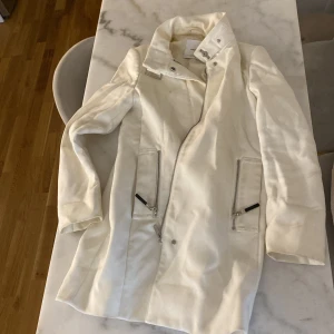 Vit rock från Mango med dragkedjor - Snygg vit rock från Mango med coola dragkedjor på fickorna och snedställd dragkedja framtill. Jackan har hög krage med tryckknapp och stilren design som passar perfekt till många outfits. Materialet känns mjukt och har en lätt glans. 