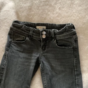 Mörkgrå jeans från Gina Tricot, stl 140 - Snygga mörkgrå jeans från Gina Tricot i klassisk femficksmodell. Byxorna har dubbla knappar i midjan, justerbar resår på insidan och bakfickor med lock och knapp. Perfekta för dig som gillar en stilren look.