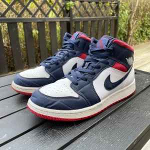 Nike Air Jordan 1 Mid USA Olympics - Snygga Nike Air Jordan 1 Mid sneakers i marinblått, vitt och rött. Klassisk siluett med snörning, röd yttersula och ikoniska Jordan Wings-loggan på sidan. Ovandel i skinn och syntet, med vadderad krage för extra komfort. Perfekta för dig som vill sticka ut med stil.