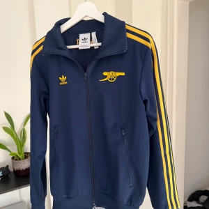 Adidas Arsenal blå track jacket - Snygg blå track jacket från Adidas x Arsenal med gula detaljer och broderad kanon på bröstet. Klassiska gula ränder längs ärmarna, dragkedja framtill och två fickor. Broderad Arsenal-text i nacken. Perfekt för dig som gillar sportig stil.Använd två gånger och tvättad en gång, OBS FINNS UTE PÅ FLER SIDOR