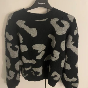Svart-grå leopardmönstrad stickad tröja - Snygg svart stickad tröja med grått leopardmönster. Tröjan har rund halsringning och långa ärmar, perfekt för dig som vill ha en trendig och mysig look. Passar till både jeans och kjol för en cool stil.