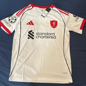 Liverpool bortatröja Mac Allister 10 - Liverpool FC bortatröja med Mac Allister 10-tryck. Vit med röda detaljer, svarta linjer och klubbmärke på bröstet. Kortärmad modell i lätt polyester med Champions League-märke på ärmen och Standard Chartered som sponsorlogga.