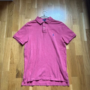 Rosa pikétröja från Ralph Lauren - Passar S. Snygg rosa pikétröja från Polo Ralph Lauren i slim fit-modell. Klassisk krage, korta ärmar och två knappar framtill. Ikoniska blå loggan broderad på bröstet.