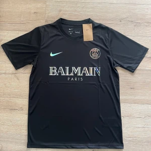 Svart PSG x Balmain fotbollströja Nike - Svart fotbollströja från Nike i samarbete med PSG och Balmain. Tröjan har korta ärmar, rund hals och är gjord i ett lätt, tekniskt Dri-FIT-material. Framsidan pryds av holografiska loggor för Nike, PSG och Balmain Paris. Perfekt för dig som gillar stilren och exklusiv sportstil.