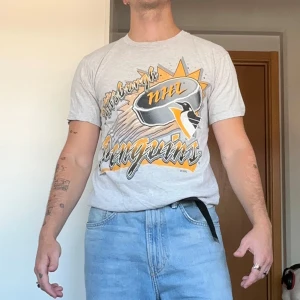 Pittsburgh Penguins vintage t-shirt - Grym vintage Pittsburgh Penguins t-shirt i ljusgrått med stor färgglad NHL-logga och tryck framtill. Klassisk passform, korta ärmar och rund hals. Tillverkad i 100% bomull, tung och skön kvalitet. Perfekt för dig som gillar sportig retrostil. Sitter som en S. Har lite små färgfläckar där nere, syns på bild 4. 