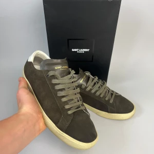 Saint Laurent Paris bruna sneakers mocka - Snygga bruna sneakers från Saint Laurent Paris i mocka med grå snörning och vit sula. Skorna har en stilren design med diskret logga i guld på plösen och vit detalj vid hälen. Perfekta för dig som gillar lyxig streetstyle.