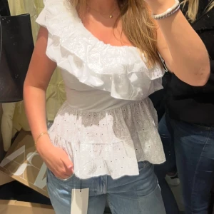 Vit omlott blus med volang - Superfin vit blus med stora volanger och omlott-skärning över ena axeln. Blusen har broderade detaljer och är ärmlös, perfekt för sommarens alla tillfällen. Snyggt fall och luftig känsla, passar till både jeans och kjol.