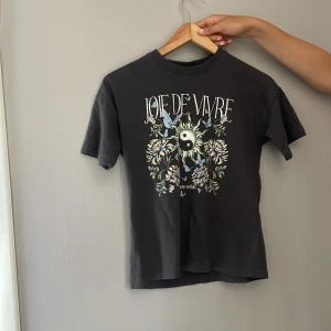 Svart t-shirt med tryck från Lindex - Cool svart t-shirt från Lindex med trycket 'Joie de Vivre' och ett stort motiv med yin-yang, blommor och fjärilar på framsidan. Det står initialer skrivna på baksidan av lappen men såklart inget som syns💕❤️