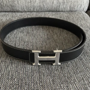 Svart Bälte med H-spänne - Snyggt svart bälte med ikoniskt H-spänne i silverfärgad metall. Bältet är gjort i äkta skinn och har vita kontrastsömmar längs kanterna. Perfekt accessoar för att lyfta din outfit med en lyxig touch.