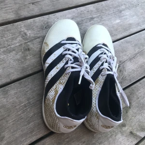 Adidas vita och svarta fotbollsskor - Snygga Adidas fotbollsskor i vitt med svarta ränder och guldiga detaljer. Skorna har snörning och platt sula, perfekt för inomhusspel. Klassisk design med tydlig Adidas-logga på hälen och sulan. Lätta och smidiga för snabba rörelser på planen.