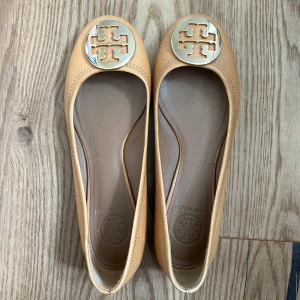 Beige ballerinaskor från Tory Burch - Snygga ballerinaskor i beiged skinn från Tory Burch med rund tå och platt sula. Skorna har en stor guldfärgad logga framtill som ger en lyxig touch. Perfekta för dig som gillar klassisk och stilren design.   De är i nyskick, knappt använda Nypris: 3200kr Mitt pris: 700kr