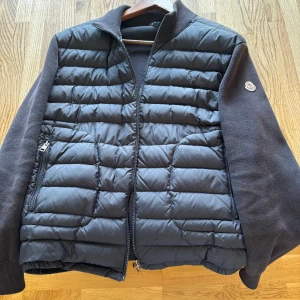 Moncler cardigan  - Snygg svart cardigan från Moncler med quiltad framsida och mjuka stickade ärmar. Cardiganen har dragkedja framtill, ståkrage och två fickor med dragkedja. Klassisk Moncler-logga på ärmen. Perfekt för lager-på-lager och streetstyle.
