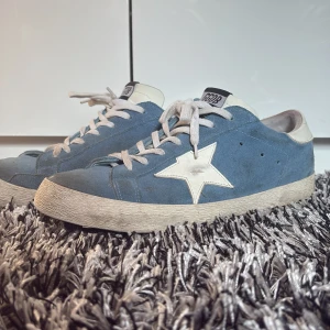 Blå mocka sneakers från Golden Goose - Snygga blå sneakers från Golden Goose med vit stjärndetalj på sidan och vita skosnören. Skorna är i mocka med vit hälkappa och platt sula. Klassisk GGDB-logga på plösen och väldigt unik. Väldigt bra skick. Storlek 43. Hör av dig!!